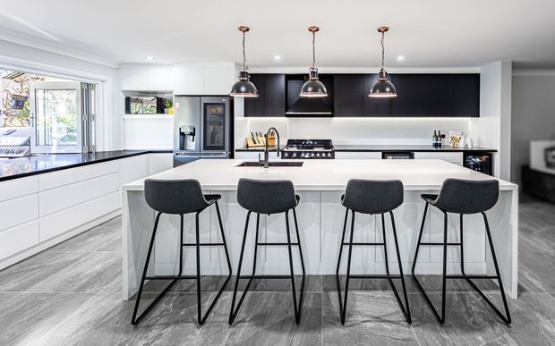 Bright contemporary kitchen centred on a large white island with seating for four. The space features extensive white cabinetry, a black upper cabinet and rangehood wall, stainless-steel appliances, a gas cooktop, pendant lights, and grey wood-look flooring. Large glazing and an open connection to an outdoor area are visible at left.