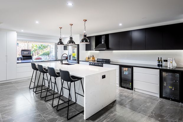 A bright, contemporary kitchen with a large white stone island, four black bar stools, matte black overhead cabinetry, integrated appliances, pendant lighting, and grey stone-look flooring. A window above the rear bench brings in natural light and shows greenery outside.