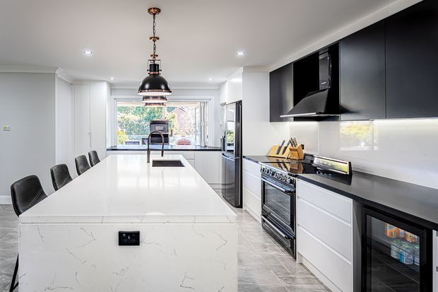 Bright contemporary kitchen with a large white stone island, integrated seating, black pendant lighting, glossy black overhead cabinetry, white lower cabinetry, stainless-steel appliances, and a dark benchtop. A dining or living area is visible beyond the kitchen.