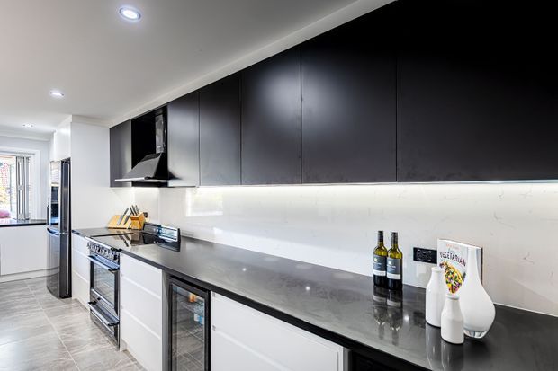 Modern galley kitchen with extensive matte black upper cabinetry, white lower drawers, a long dark stone-look benchtop, and a bright white marble-look splashback illuminated by under-cabinet lighting. Stainless-steel appliances, a black rangehood, integrated refrigeration, glass-front beverage storage, and pale tiled flooring complete the space.
