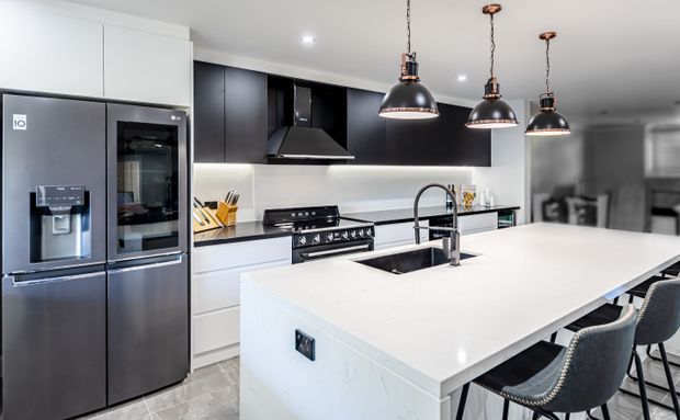 Contemporary kitchen featuring a large white stone island with integrated sink and high-arched tap, seating for several people, dark cabinetry, a stainless-steel refrigerator and oven, a freestanding range, and three black pendant lights. The palette is predominantly white, black, and grey with a polished, clean-lined finish.