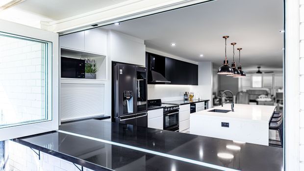 A bright contemporary kitchen viewed through a wide opening, featuring glossy black and white cabinetry, a large white island with integrated sink, black stone-look benchtops, stainless-steel appliances, and three black pendant lights. Dining and living areas are visible beyond the kitchen.