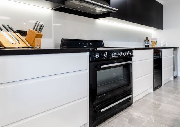 Contemporary kitchen featuring sleek white handleless cabinetry, a black SMEG freestanding range cooker, black overhead cabinetry, integrated under-cabinet lighting, a pale stone-look splashback, and light grey tiled flooring. Timber knife blocks and small countertop items provide limited warmth against the monochrome fit-out.