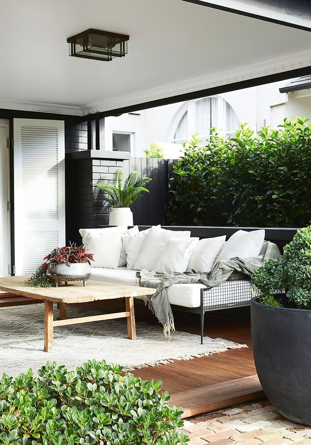 A covered outdoor courtyard or terrace styled as a comfortable lounge, with a white sectional sofa, rustic timber coffee table, patterned rug, abundant potted greenery, black-painted brick walls, dark timber decking, and a ceiling-mounted lantern. The space opens toward a planted courtyard and includes a bright arched window beyond.
