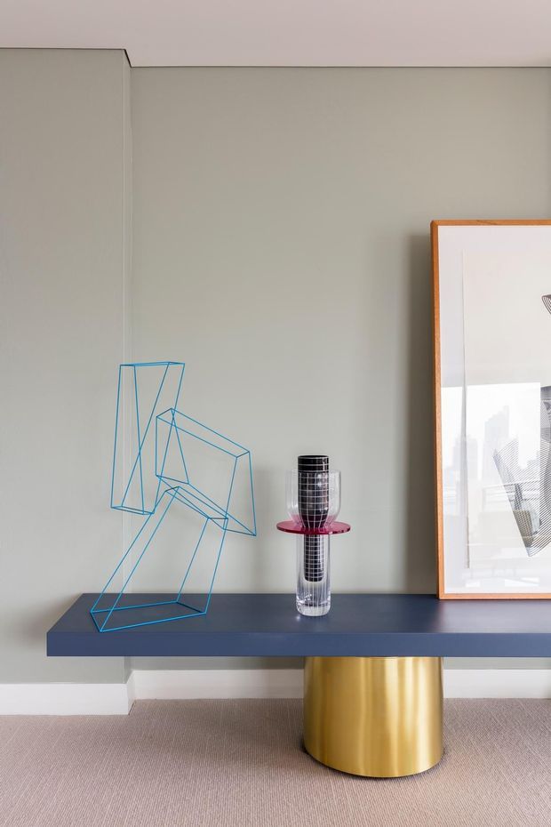 A minimalist interior vignette featuring a long dark blue console table with a cylindrical brushed gold pedestal base. The console is styled with a geometric blue wire sculpture and a tall reflective glass-and-metal vase, alongside a large framed artwork. The pale grey wall, light carpet, and restrained decorative styling create a refined contemporary look.