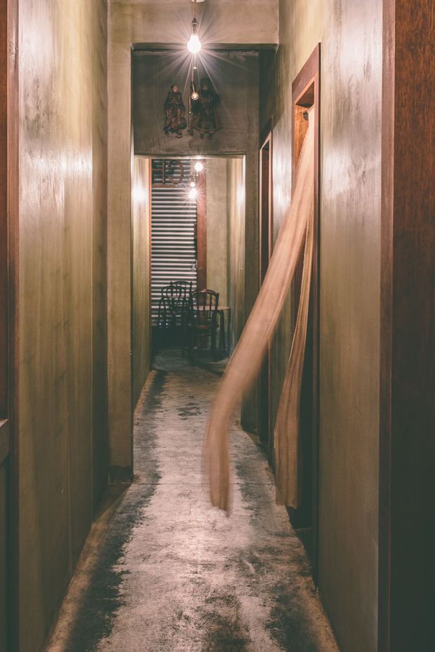 A narrow, atmospheric interior corridor with dark timber wall panelling, a raw mottled concrete floor, warm pendant lights, and a sculptural curved timber handrail or feature element running along the right side. The corridor leads toward a small seating area visible at the far end, with reflective surfaces creating depth. The mood is moody, rustic, and industrial-inspired.