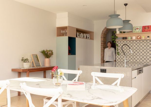 Bright open-plan kitchen and dining area with a large white marble-look dining table, white cross-back chairs, pale cabinetry, patterned tile splashback, arched exposed-brick opening, and pastel pendant lights. Decorative shelving and a sideboard add a warm, playful character to the space.
