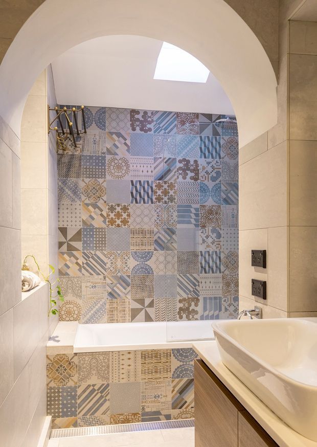 A compact bathroom viewed through a large curved archway. The bath enclosure is finished with a patchwork feature wall and bath-front of patterned blue, grey, beige and white tiles, while the surrounding walls use large beige tiles. A white vessel basin sits on a timber-toned vanity, with a wall-mounted shower fixture, skylight and dark electrical fittings visible.