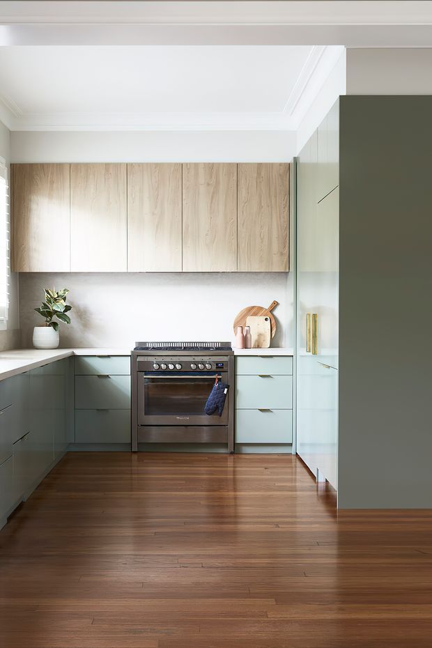 A bright, contemporary U-shaped kitchen with pale timber overhead cabinetry, muted sage-green lower cabinets, a stainless-steel freestanding oven, and integrated tall storage. The kitchen features a light stone benchtop and splashback, warm timber flooring, white walls with simple cornices, and understated brass-toned handles. Natural light enters from a side window, creating a clean, refined atmosphere.