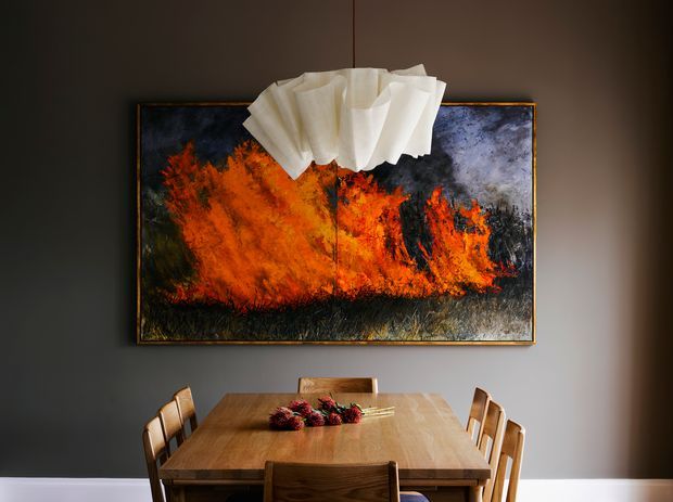 A moody dining room with a large rectangular timber dining table and matching wooden chairs, set against a dark grey wall. A vivid abstract artwork in orange, black, and white hangs above the table, while a sculptural white pendant light is suspended centrally overhead. The room has a warm, dramatic and contemporary feel.