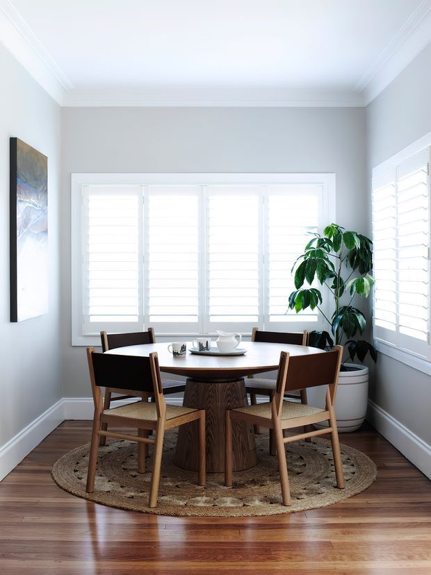 Bright, compact dining area with a round mid-century-inspired wooden dining table and four wooden chairs on a woven round rug. Pale grey walls, white trim and plantation shutters surround the room, with a large potted plant and artwork adding natural and decorative accents. Polished timber flooring and abundant filtered daylight create a warm, airy atmosphere.