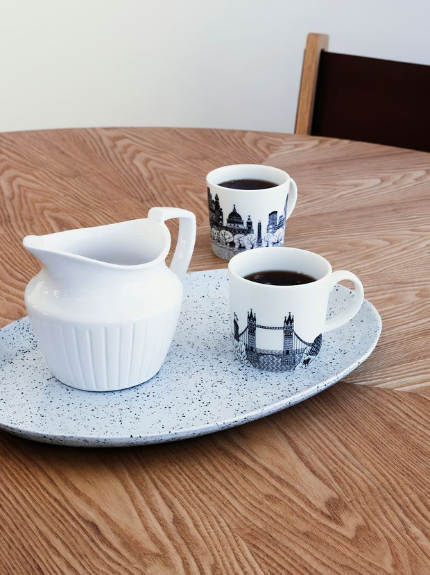 Close-up still life of a tea setting on a light wood dining table, featuring a white ceramic milk jug and two illustrated teacups on a speckled pale tray. A dark wood dining chair is partially visible in the background.
