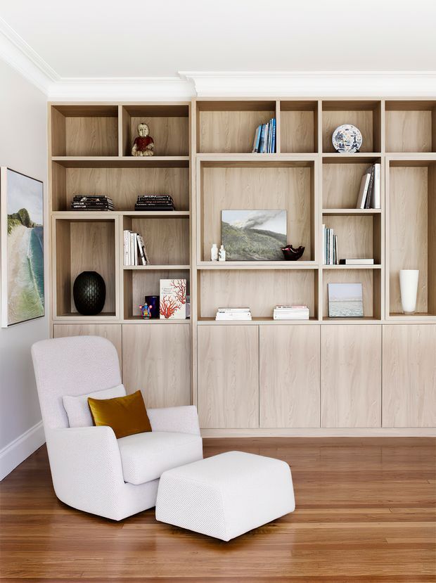A refined living room with custom full-height timber built-in shelving and closed cabinetry, displaying books and decorative objects. A white upholstered lounge chair with a mustard cushion and matching ottoman sits on warm timber flooring. The room features white walls, detailed crown moulding, framed artwork, and a calm, tailored aesthetic.