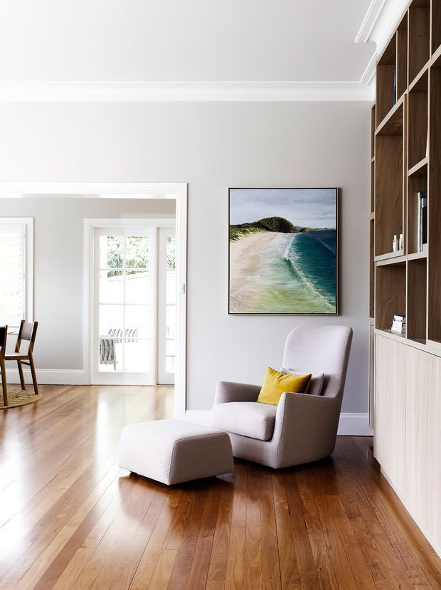 Bright, refined living room with polished timber flooring, pale grey walls, white decorative ceiling cornices and trims, and a built-in timber display wall with white lower cabinetry. A sculptural upholstered armchair and matching ottoman sit beside a framed coastal artwork, with a doorway opening toward a brighter adjoining space.