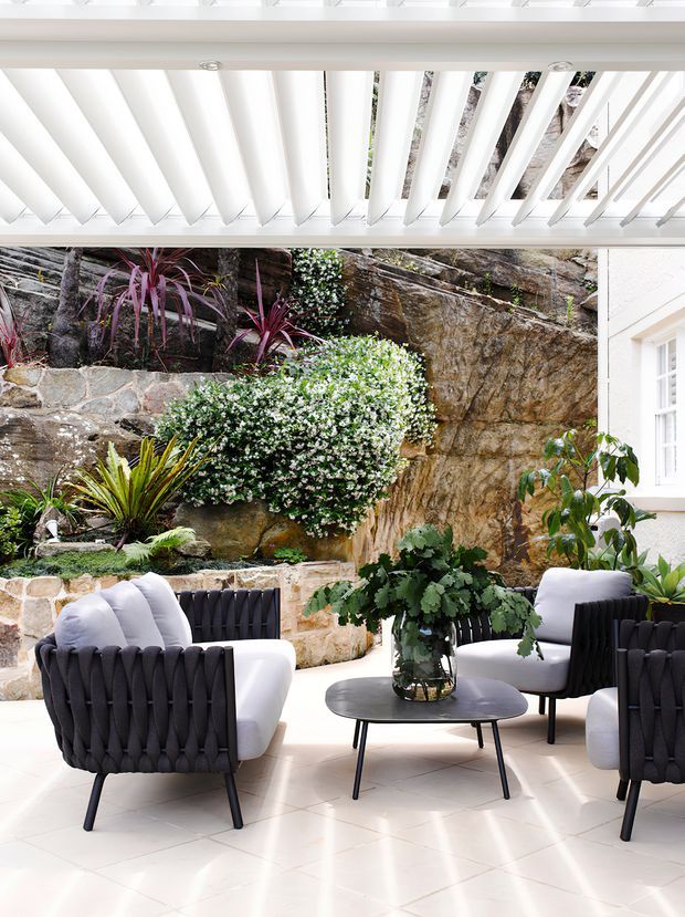 A bright, sheltered outdoor lounge or courtyard with white-painted overhead louvres, pale tiled flooring, dark woven lounge chairs with white cushions, and a round black coffee table. A natural stone retaining wall with dense ornamental planting forms the backdrop, while a white-framed window connects the space to the building.