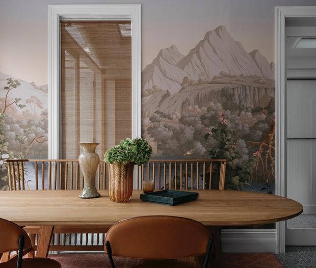 A refined dining room with a large oval timber dining table and leather dining chairs. The walls feature a dramatic scenic mural depicting mountains, trees, and foliage, complemented by white decorative trim and a window with timber blinds. A vase and floral arrangement sit on the table, creating a warm, richly layered interior.