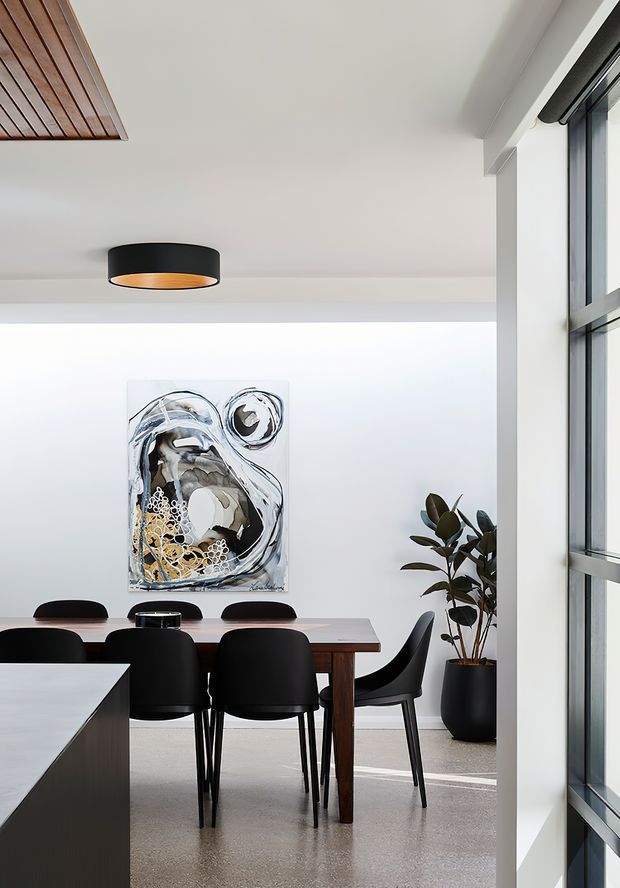 Bright contemporary dining area with a long dark timber dining table, black dining chairs, abstract wall artwork, a large potted plant, polished concrete flooring, white walls and ceiling, and expansive black-framed windows. A black drum pendant light and timber ceiling detail add contrast.