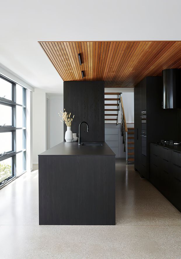 A refined contemporary kitchen with a large dark timber island, integrated black cabinetry, matte black tapware and appliances, and a warm linear timber-slat ceiling. A compact open stair with timber treads is visible beyond the kitchen. Full-height windows bring in abundant natural light, while the polished concrete or terrazzo-like floor reinforces the minimalist, high-contrast scheme.