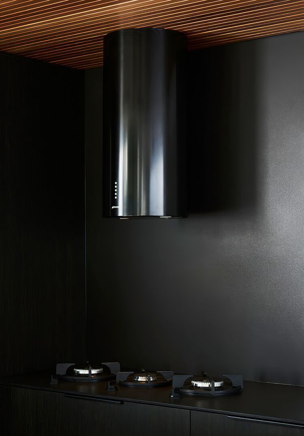 A dramatic close-up of a dark, contemporary kitchen featuring a three-burner gas cooktop set into black cabinetry and a large cylindrical stainless-steel rangehood. The space has a matte charcoal wall splashback and a striking linear timber-slat ceiling with warm recessed accents.