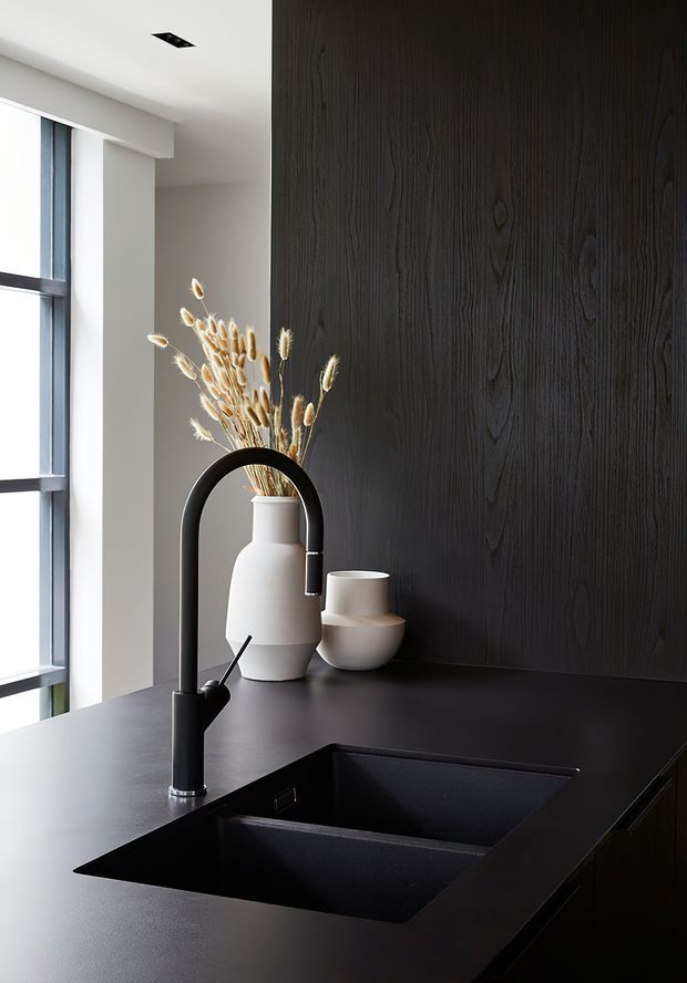 A refined, minimalist kitchen vignette featuring a matte-black double-bowl undermount sink and sculptural black gooseneck faucet set into a dark benchtop. A dark vertical timber-grain feature wall contrasts with the white wall and tall black-framed window. White ceramic vessels with dried grasses add a restrained decorative accent.