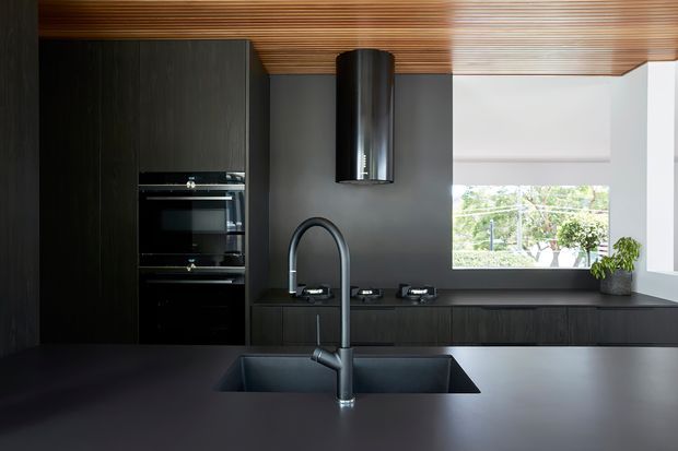 A contemporary dark-toned kitchen with full-height black cabinetry, integrated double ovens, a large island with a deep undermount sink and sculptural black tap, and a black cooktop wall beneath a cylindrical rangehood. A warm timber slatted ceiling contrasts with the charcoal joinery and benchtops, while a large window brings in daylight and views of greenery.