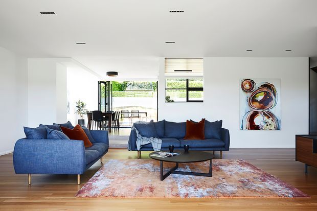 Bright, open-plan contemporary living room with white walls and ceiling, timber flooring, blue upholstered seating, a dark oval coffee table, and a large patterned area rug. A dining area and garden are visible beyond the living space, with abstract artwork and streamlined recessed lighting adding visual interest.