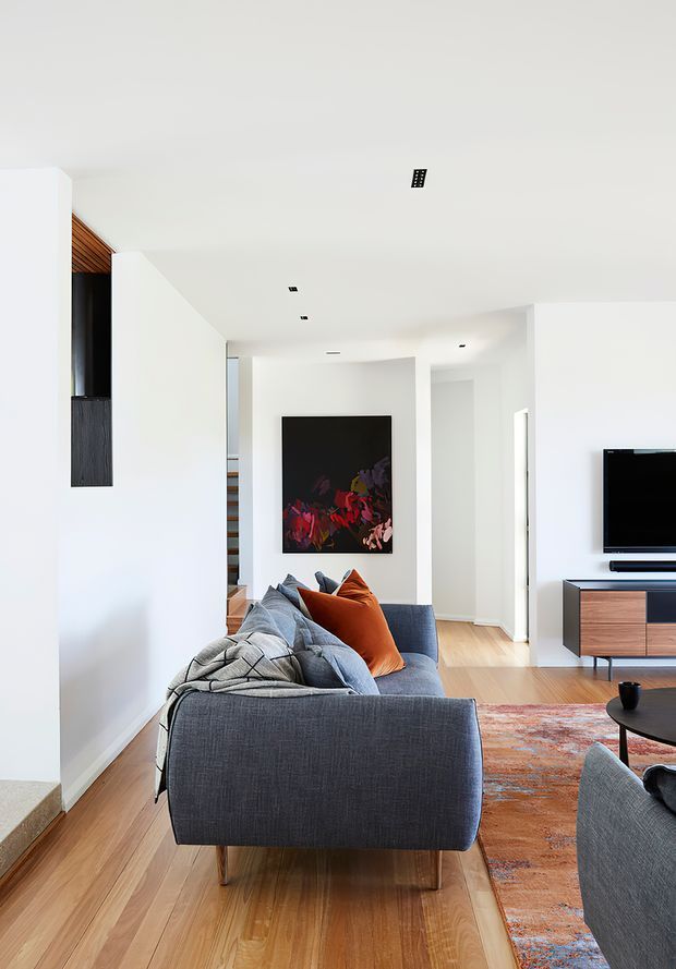 Bright contemporary living room with white walls and ceiling, warm timber flooring, a large grey sofa with orange and blue cushions, a black-framed artwork, and a television on a timber media console. The room has recessed lighting, clean lines, and an open connection toward a hallway and stair area.