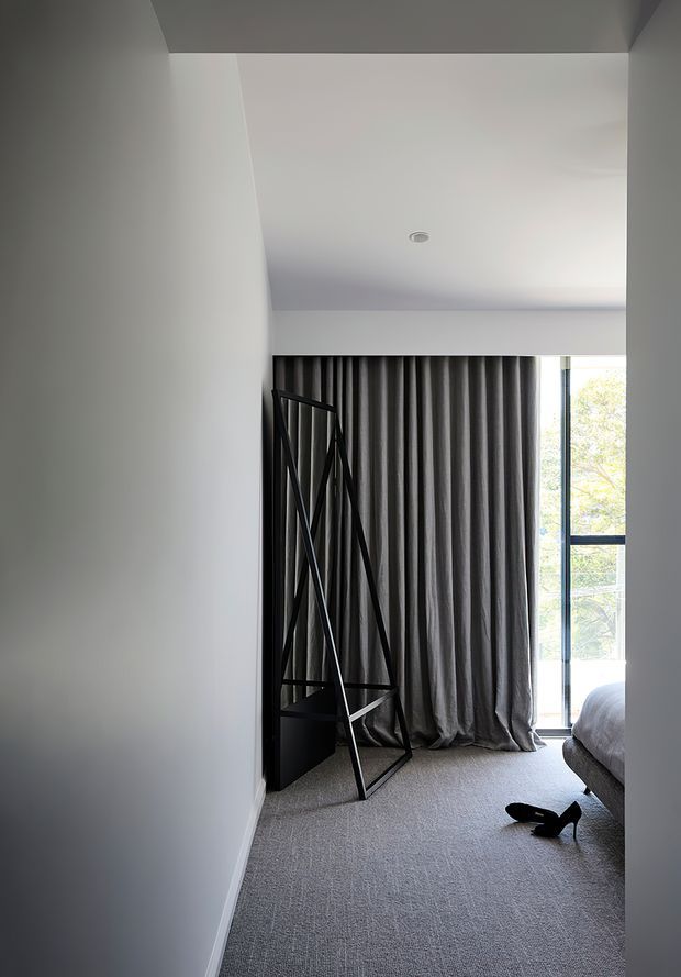A minimalist bedroom interior viewed from the entry, with white walls and ceiling, grey carpet, full-height dark grey curtains, and a large window overlooking trees. A portion of the bed is visible at right, alongside a black angular freestanding clothes rack or sculptural storage frame.
