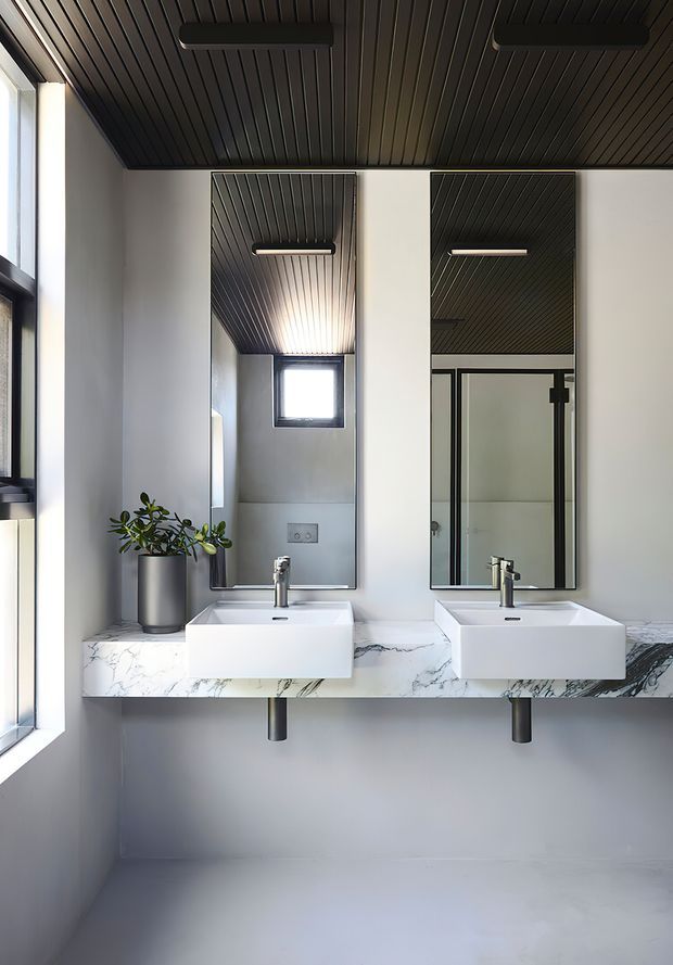 Contemporary double vanity bathroom with two wall-mounted white rectangular basins, a white marble-look countertop, tall mirrors, dark slatted ceiling, black-framed glazing, and minimalist black fixtures. A small potted plant adds a natural accent.