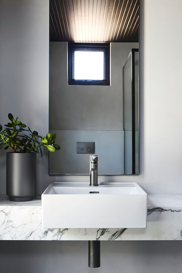 A close-up view of a contemporary bathroom vanity. A white vessel basin sits on a dramatic white marble benchtop with grey veining, paired with a chrome tap and black-framed rectangular mirror. The bathroom has pale grey walls, a dark linear-panel ceiling, integrated wall-hung cistern controls, and a potted plant.