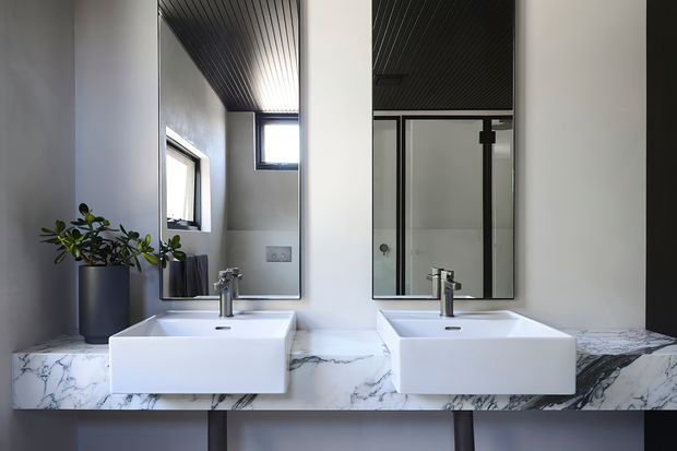 Contemporary bathroom vanity with two white rectangular basins set on a dramatic white marble-effect stone benchtop, beneath twin mirrors. Dark framed shower glazing, charcoal slatted ceiling, grey walls, chrome tapware, and a potted plant create a refined monochrome scheme.