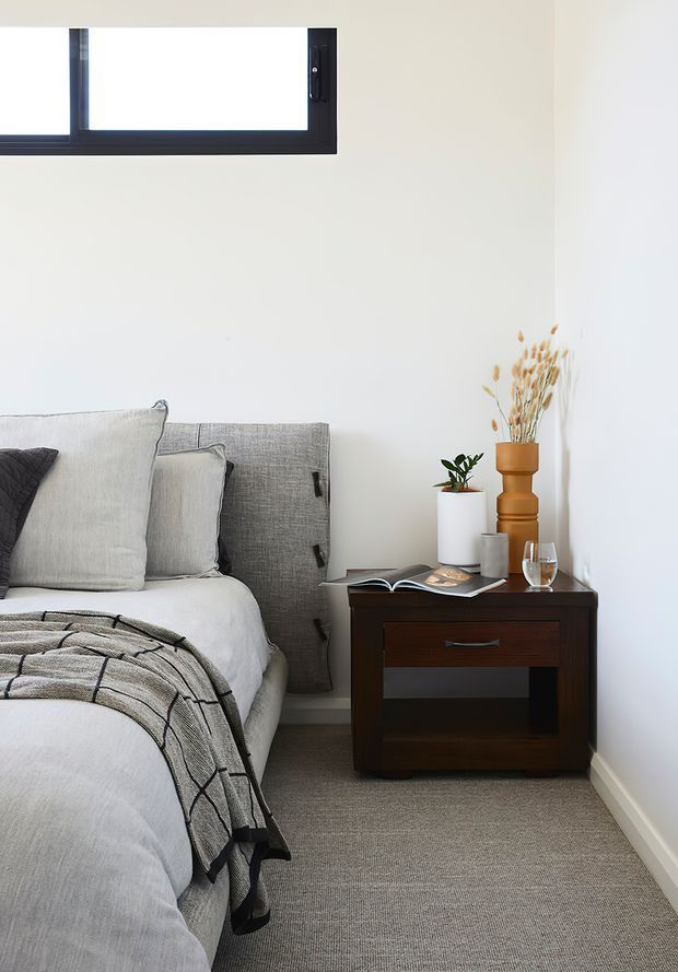 A calm, minimalist bedroom corner with a neatly made bed, layered pale grey and white bedding, a textured upholstered headboard, and a dark timber bedside table. A small plant, dried grasses, a glass, and an open magazine sit on the table. White walls, grey carpet, and a high horizontal black-framed window create a bright, understated interior.