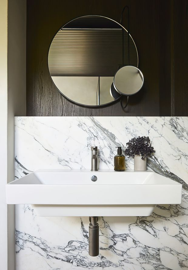 A compact, sophisticated bathroom vanity featuring a wall-mounted white rectangular basin with integrated tap, set against dramatic white marble-look veined wall panels. The upper wall is clad in dark vertical timber, with a large round mirror and smaller circular magnifying mirror. Minimal toiletries and a small plant sit on the basin, creating a refined, moody look.