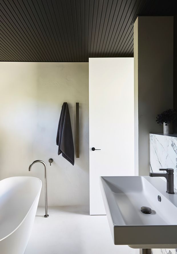 A minimalist contemporary bathroom featuring a freestanding white bathtub, wall-mounted towel rail, sculptural basin with dark faucet, white marble-look splashback, and a dark linear-panel ceiling. The space has a restrained monochromatic palette with warm natural light and clean geometric forms.