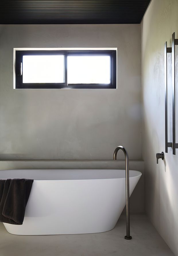 A minimalist bathroom featuring a freestanding white bathtub, slim floor-mounted brushed metal bath filler, dark-framed horizontal window, pale plastered walls, and a dark linear slatted ceiling. A dark towel hangs over the tub, while a wall-mounted mirror and fittings are partially visible at right.