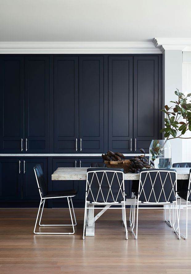 A refined dining area within an open-plan kitchen, featuring full-height dark navy paneled cabinetry with white crown moulding, a pale stone dining table, white geometric wire-frame chairs with dark seats, and warm timber flooring. A leafy plant and glass vase provide natural accents. The space has a strong, high-contrast classic-contemporary character.