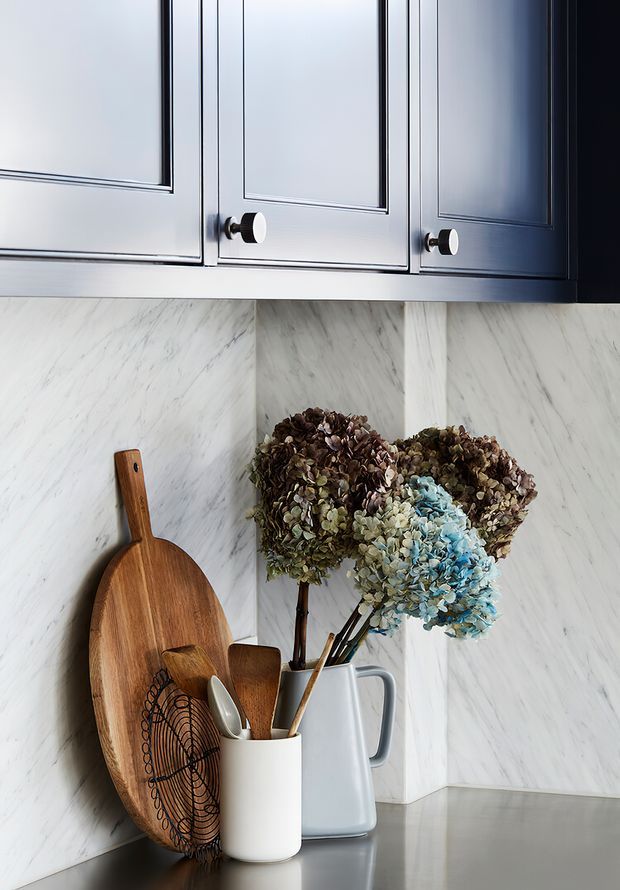 Close-up detail of a refined kitchen corner with dark blue-grey framed upper cabinetry, white marble-look backsplash, and a grey countertop. Wooden serving boards, utensils, ceramic vessels, and dried hydrangeas provide warm decorative accents.