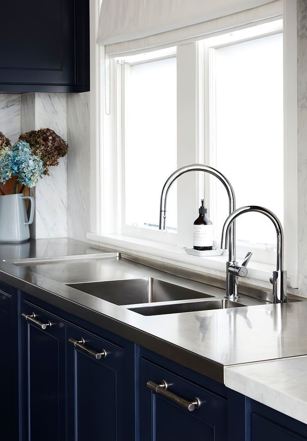 Close-up view of a refined kitchen sink zone beneath bright windows, featuring deep navy shaker-style cabinetry, a polished stainless-steel benchtop and double-bowl undermount sink, paired with chrome gooseneck and secondary taps. White marble-look wall and counter surfaces, a roller blind, and understated styling complete the light, classic composition.