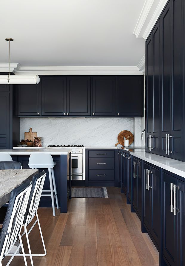 A refined galley-style kitchen with deep navy-blue shaker cabinetry, extensive white marble benchtops and splashback, stainless-steel appliances, and polished chrome hardware. A large stone-topped island with white bar stools occupies the foreground, while timber flooring, white crown moulding, and a linear pendant light add warmth and classic detailing.