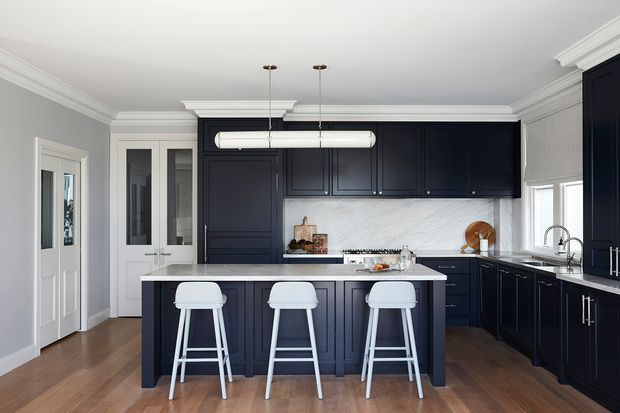 A refined, spacious kitchen with dark navy-blue shaker cabinetry, a large central island with seating for three, white marble-look splashback, light stone benchtops, timber flooring, and white decorative ceiling moulding. Tall windows and glass-panelled doors bring in natural light, while a linear pendant adds a contemporary touch.
