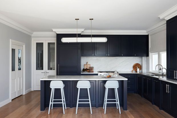 A refined traditional kitchen with deep navy-blue paneled cabinetry, a large central island with three white bar stools, a white marble-look backsplash and benchtop, timber flooring, white walls, and decorative crown moulding. Brass-toned pendant fittings and stainless fixtures provide subtle contrast. The room is bright, orderly, and spacious.
