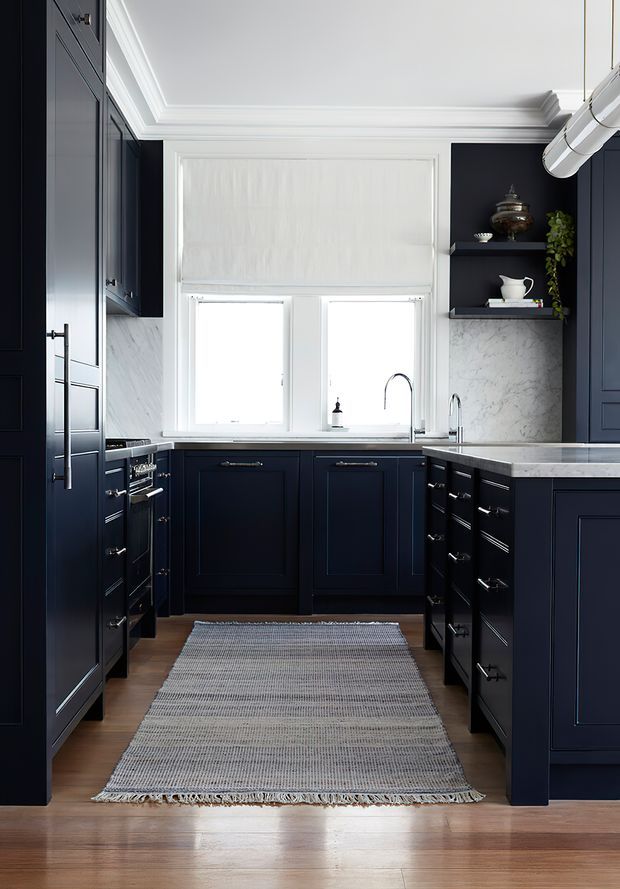 A compact, highly finished galley kitchen with deep navy-blue shaker cabinetry, white marble-look splashback and benchtops, polished chrome hardware and tapware, and warm timber flooring. A large window provides strong natural light, while open shelving, integrated appliances, and a pale woven runner complete the refined traditional interior.