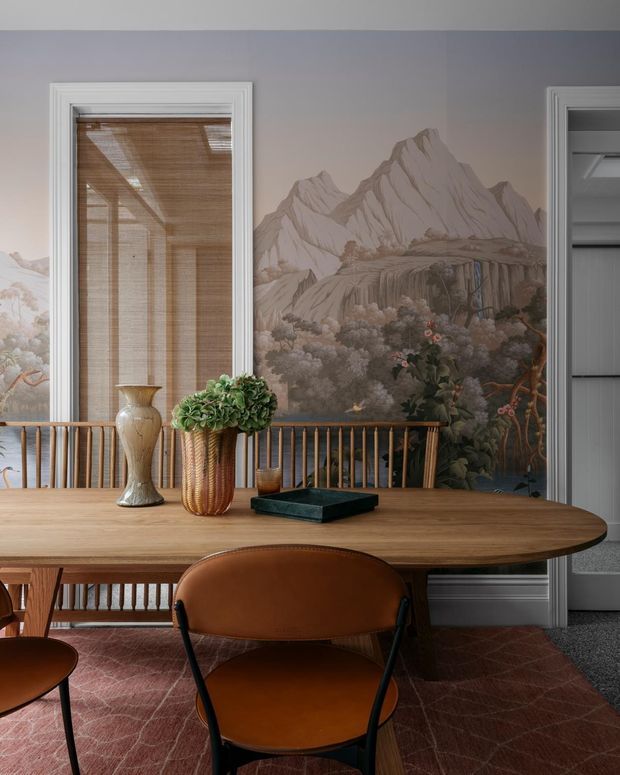 A refined dining room with a large oval timber dining table, warm brown leather chairs, a woven pendant or blind visible through the window, and a dramatic floor-to-ceiling botanical mountain mural. White decorative wall and door trims, a timber balustrade, layered greenery, and a muted red patterned rug create a richly detailed, warm interior.