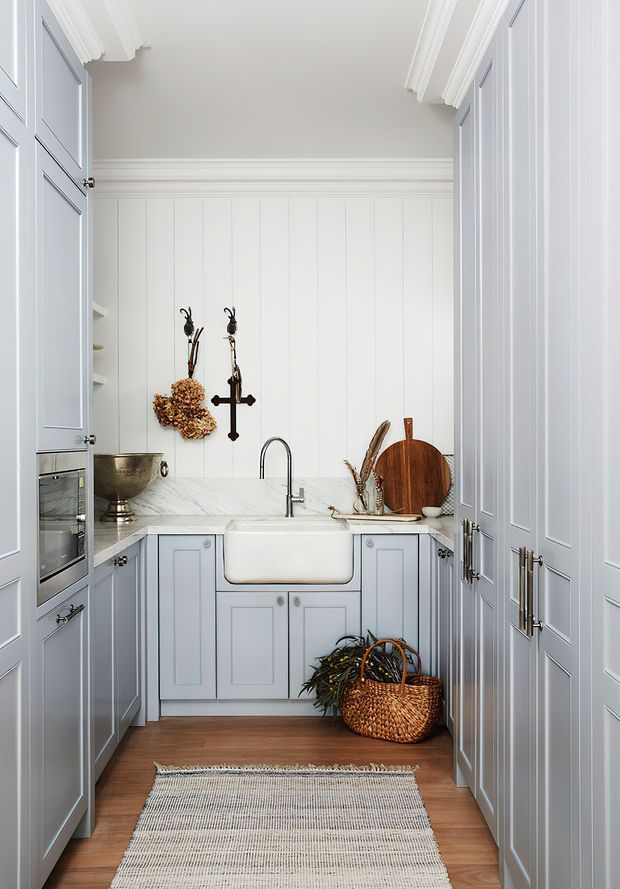 A compact, highly organized butler's pantry or scullery with floor-to-ceiling pale blue-grey panelled cabinetry, a white marble benchtop, farmhouse-style apron-front sink, integrated oven, open shelving, and brushed metal hardware. White vertical wall panelling and detailed crown moulding create a refined traditional character, while timber flooring, woven rug, cutting boards, dried botanicals, and baskets add warmth.