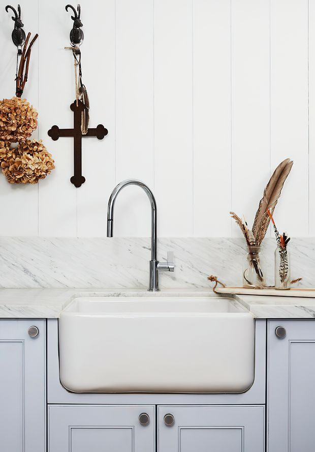 Close-up interior view of a refined kitchen sink area featuring a deep white farmhouse apron-front sink, polished chrome gooseneck tap, white marble benchtops and splashback, pale blue-grey framed cabinetry, and vertically panelled white walls. Dried hydrangeas, feathers, and a dark wooden cross provide decorative accents.