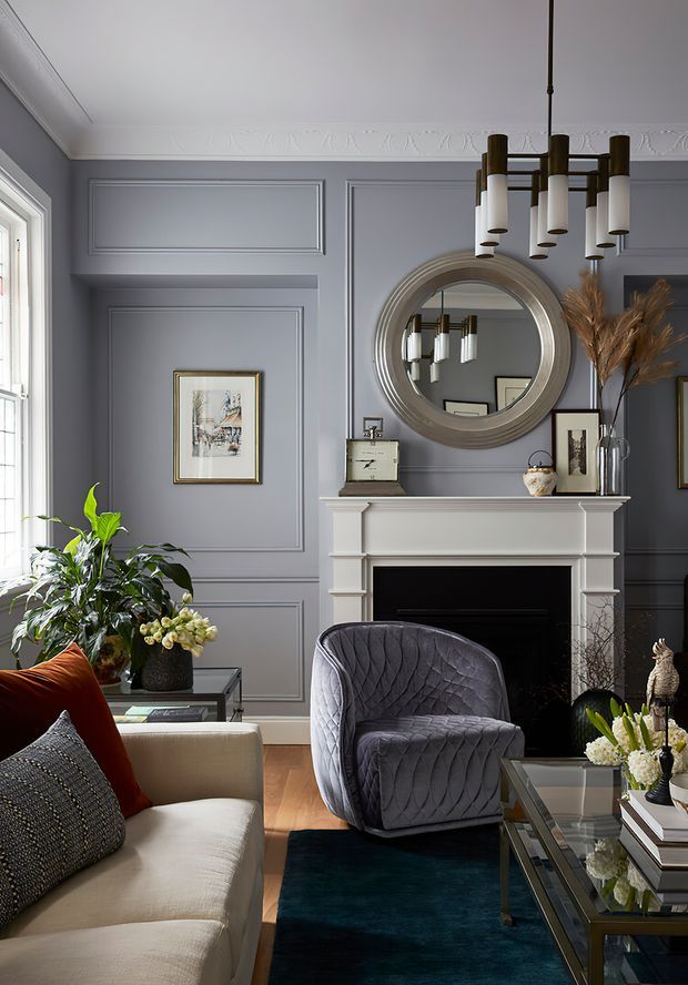 Elegant living room with dark grey-blue decorative wall panelling, ornate ceiling cornice, a classical mantel fireplace, large round mirror, layered artwork, and a sculptural modern chandelier. A cream sofa, quilted grey armchair, glass coffee table, indoor plants, and warm timber flooring create a refined, richly layered interior.