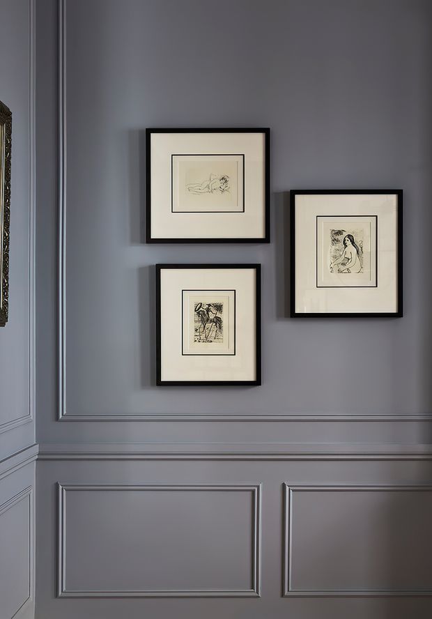 A refined wall vignette featuring three black-framed monochrome artworks arranged on a muted grey-blue wall. The wall has substantial decorative raised-panel wainscoting and moulded trim, creating a formal, classic interior character.