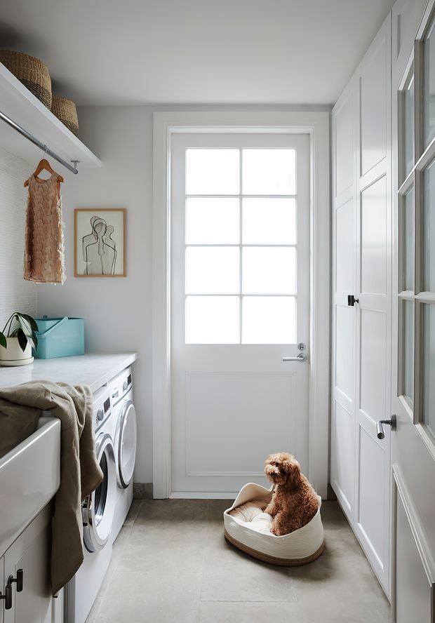 Bright, compact laundry room with a front-loading washer and dryer beneath a pale stone-look benchtop, a deep utility sink, open hanging rail, woven storage baskets, white paneled cabinetry, and a glazed exterior door. The room has soft grey-white finishes, a light tiled floor, and a small pet bed near the door.