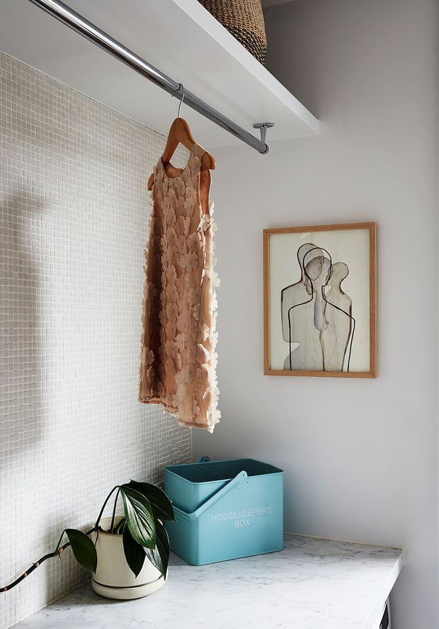 A styled laundry or utility area with a white marble-look benchtop, pale small-format mosaic tiled wall, chrome hanging rail, and a blush patterned garment hanging from a wooden hanger. A turquoise storage box and potted plant sit on the counter, with framed minimalist line artwork on the adjacent light wall and woven storage visible above.