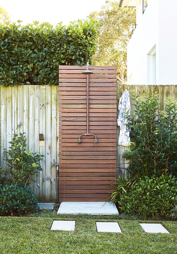 A compact outdoor shower set within a landscaped garden courtyard. The shower fixture is mounted on a tall horizontal timber slat privacy screen, with a concrete shower pad, stepping stones, timber fencing, dense hedging and shrubs surrounding it. The scene is bright and natural, with a clean contemporary garden design.